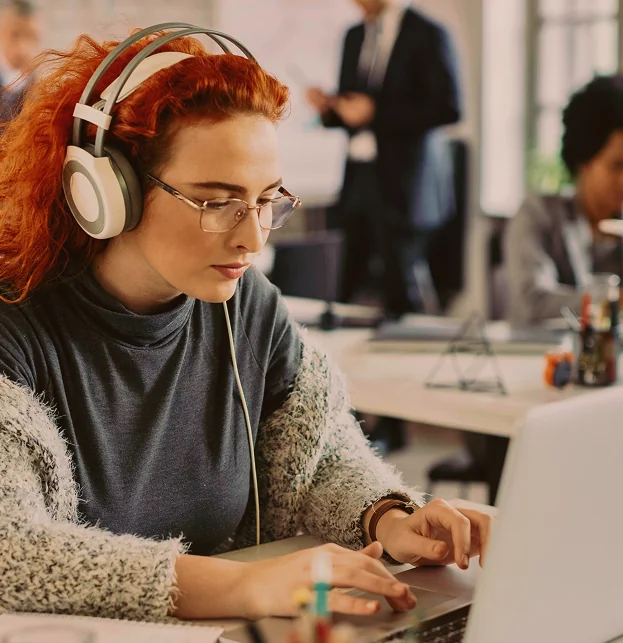 Woman with red hair wearing headphones, typing on a laptop
