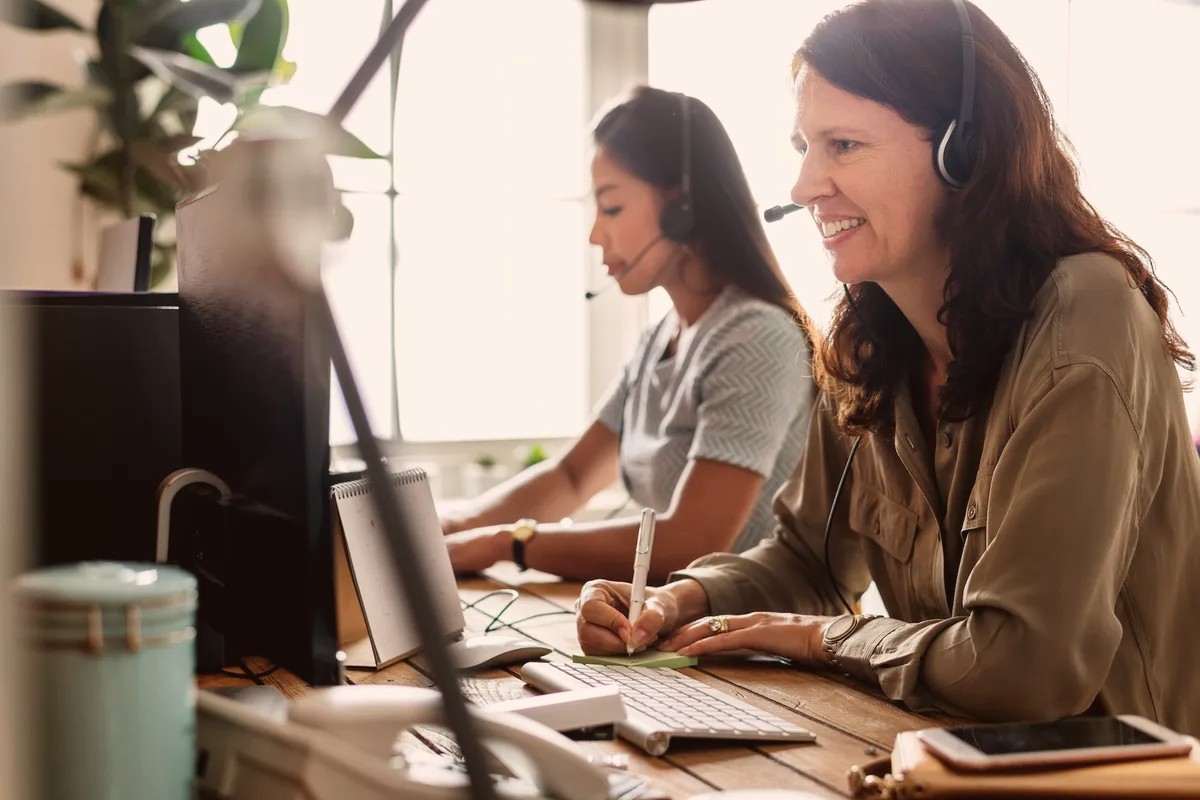Two women wearing headsets working at computers, one smiling