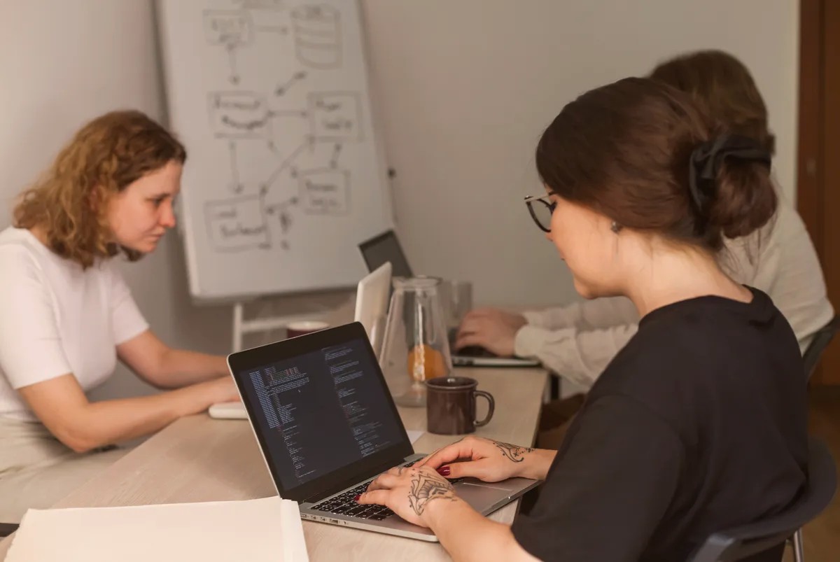 Two women working on laptops at a table, a whiteboard behind them