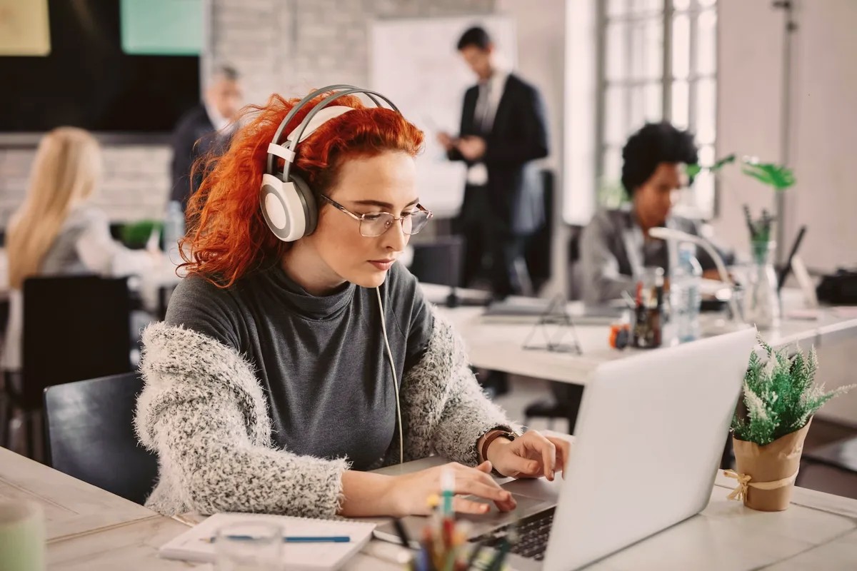Woman with red hair wearing headphones, typing on a laptop in an office