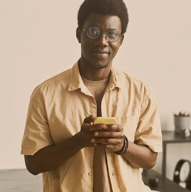 Man in glasses and a tan shirt holding a smartphone, standing in a room
