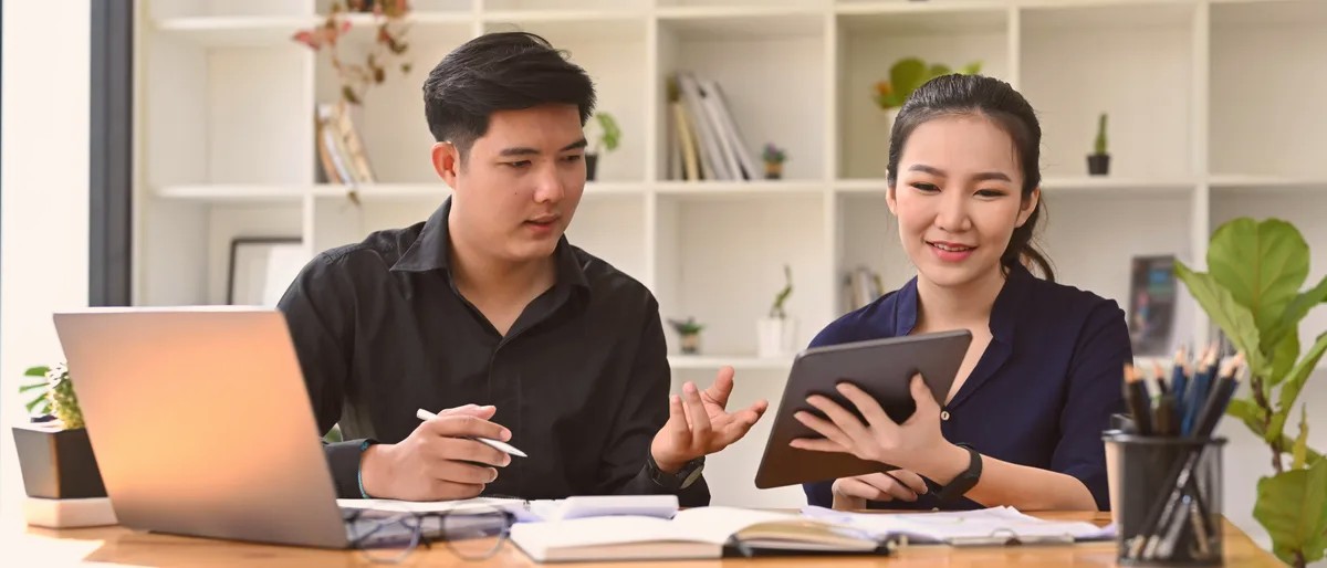 A man and a woman talking at a desk with a laptop and a tablet