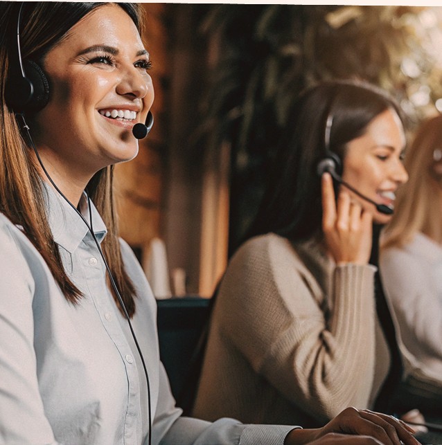 Two women wearing headsets, one smiling, at their workstations