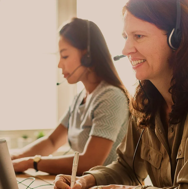 Two women wearing headsets at their desks, one taking notes