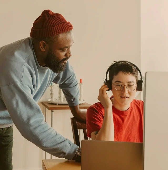 Man leaning over to help a young colleague who wears headphones at a computer