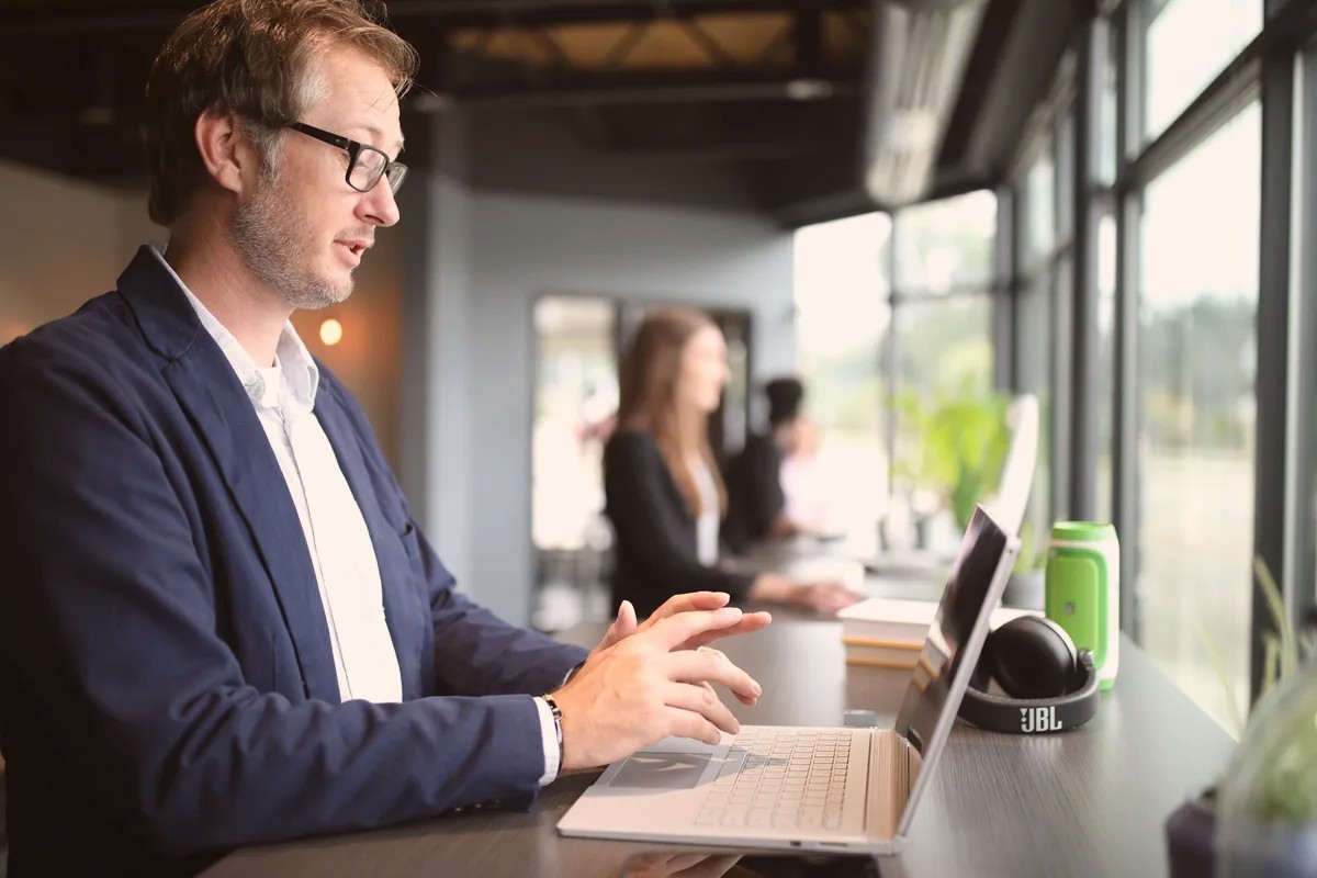 Man in a blazer typing on a laptop at a desk by a window