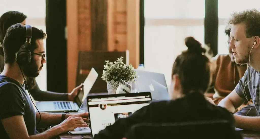 A team working on laptops around a shared table, one wearing headphones